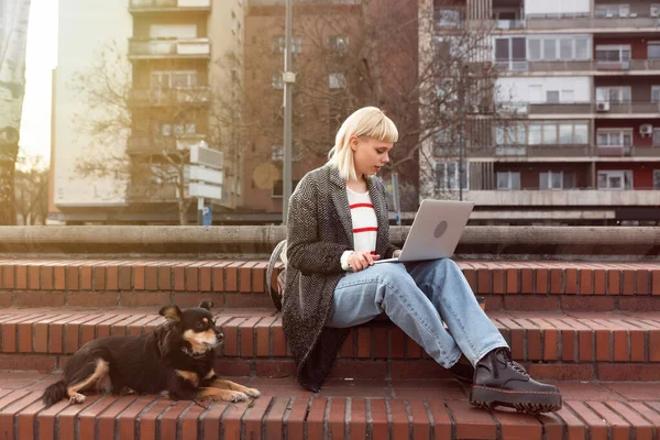 Young freelance businesswoman working outside on a laptop computer with takeaway coffee and her dog she adopted from a dog shelter. Web developer and graphic designer works out of the office
