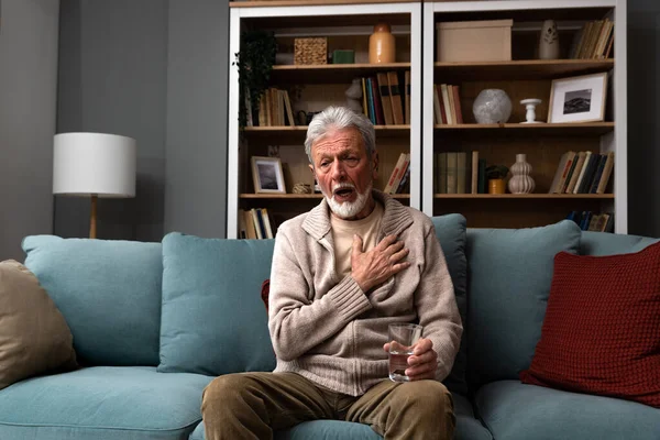 stock image Senior older man getting chest pain in living room, heart attack holding his hand on the chest sitting on sofa at home. Health care older people issue