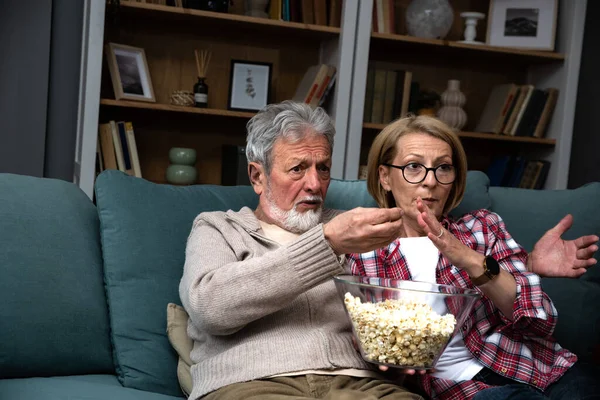 stock image Mature married couple entertaining observing terrified TV program. Senior husband and wife watching horror movie or film at home eating popcorn and making scary facial expressions