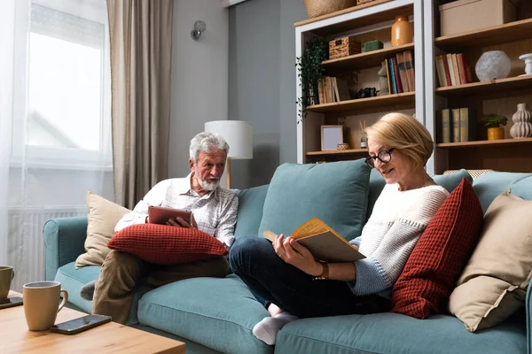 Simple living. Happy middle-aged senior couple 70s husband and wife sit relax on couch in living room reading book drinking tea together, calm elderly 60s couple booklovers rest at home enjoy life