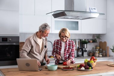 Mutlu orta yaşlı çift dizüstü bilgisayar kullanıyor. Birlikte sağlıklı beslenme diyeti salatası hazırlıyorlar. Tarifleri araştırıyorlar, internetten alışveriş yapıyorlar..