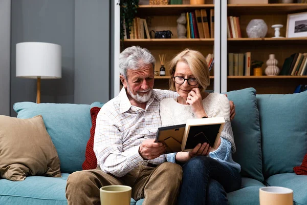 stock image Two elderly people senior couple man and woman looking at a family photo smile and hug while sitting at home on sofa. memories and nostalgia for children went abroad for better life with their family