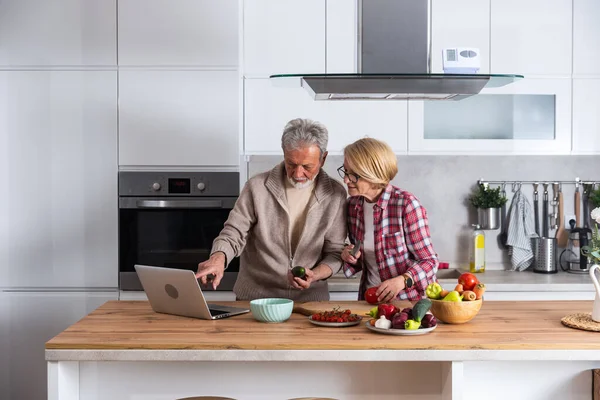 stock image Happy old middle aged senior couple using laptop computer preparing healthy food diet vegetable salad at home together, searching recipes, ordering shopping online, watching cooking class in kitchen.