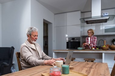 Mutlu yaşlı koca uzaktan çalışıyor ya da ev ofisinden bilgisayarla uzaktan öğreniyor. Bu arada yaşlı karısı mutfakta kahvaltı hazırlıyor. Evde orta yaşlı bir çift var..