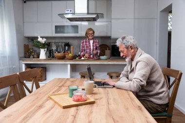 Mutlu yaşlı koca uzaktan çalışıyor ya da ev ofisinden bilgisayarla uzaktan öğreniyor. Bu arada yaşlı karısı mutfakta kahvaltı hazırlıyor. Evde orta yaşlı bir çift var..