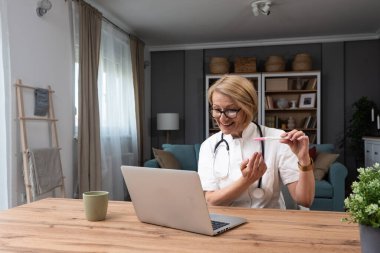 Üst düzey kadın doktor bilgisayardaki video görüntülerinde hamilelik testi gösteriyor. Ofiste steteskoplu beyaz önlüklü özel muayenehanesinde otururken.