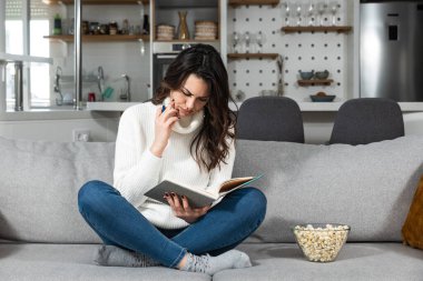 Young beautiful girl writing a love letter to her boyfriend or writing in the diary while she is smiling and sitting on the sofa in her room. Successful female having business ideas.