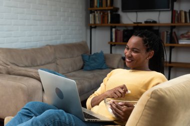 Happy african american woman streaming online movies on a laptop while snacking on popcorn and relaxing on a sofa at home. Black female enjoying a comedy, eating, laughing and having fun on a weekend