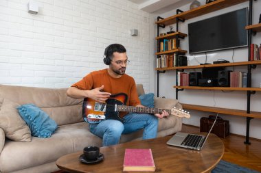 Genç erkek müzisyen kulaklık takıyor gitar çalmayı öğreniyor ve web video kursunda dizüstü bilgisayar kullanarak müzik kaydediyor. Konsantre olmuş milenyum adamı dijital stüdyo teçhizatı kullanarak yeni bir şarkı yazıyor.