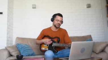 Genç erkek müzisyen kulaklık takıyor gitar çalmayı öğreniyor ve web video kursunda dizüstü bilgisayar kullanarak müzik kaydediyor. Konsantre olmuş milenyum adamı dijital stüdyo teçhizatı kullanarak yeni bir şarkı yazıyor.