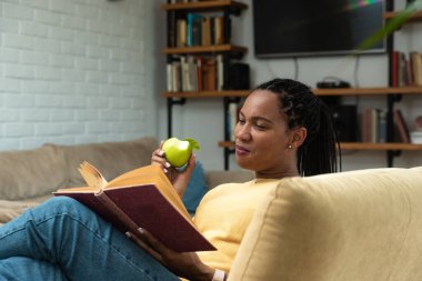 Afro-Amerikalı kadın kitap okuyor ve oturma odasında elma yiyor. Evde boş vakitlerde sağlıklı bir yaşam ve keyif..