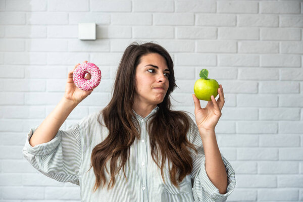 Молодая студентка сомневается в том, что выбрать здоровую пищу или сладости нездоровую пищу, держа зеленое яблоко и пончик в руках, стоящих у нее дома в гостиной. Концепция трудного выбора