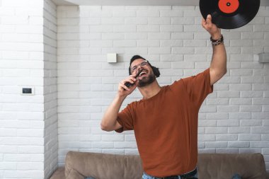 Evde şarkı söyleyen ve dans eden genç adam telefonunda şarkı söyleyen kablosuz kulaklıkla elinde LP plağıyla müzik dinliyor. Hipster müzik aşığı eski kayıt sesini dijital ortama aktardı ve eğleniyor