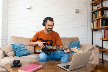 Genç adam müzik öğretmeni evdeki dizüstü bilgisayardan öğrencilere gitar dersleri veriyor. Elektro gitar çalmayı çevrimiçi kurs ve kablosuz kulaklıkla melodiyi dinlemeyi öğrenmek.