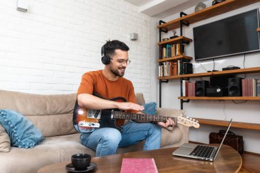 Genç adam müzik öğretmeni evdeki dizüstü bilgisayardan öğrencilere gitar dersleri veriyor. Elektro gitar çalmayı çevrimiçi kurs ve kablosuz kulaklıkla melodiyi dinlemeyi öğrenmek.