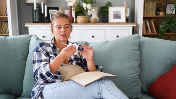 年轻美丽的女人一边在她的现代小公寓里看书 一边擦拭和擦拭她的阅读眼镜 坐在沙发上 — 图库视频影像