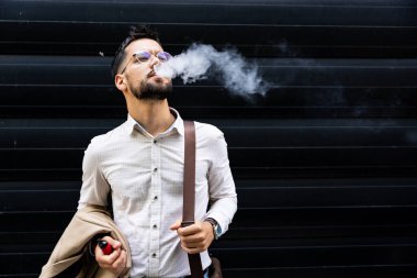 Genç işadamı, bir iş arkadaşını beklerken bir ofis binasının önünde elektronik sigara içmeye gitti.
