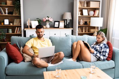 Evdeki oturma odasındaki kanepede dizüstü bilgisayarı ve kitabı olan bir kadının yanında çalışan mutlu genç adam. Serbest çalışan erkek, bilgisayar kadını üzerinde çalışıyor ve roman okuyup dinleniyor..