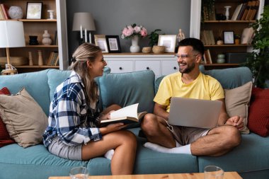Evdeki oturma odasındaki kanepede dizüstü bilgisayarı ve kitabı olan bir kadının yanında çalışan mutlu genç adam. Serbest çalışan erkek, bilgisayar kadını üzerinde çalışıyor ve roman okuyup dinleniyor..