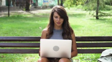 İnternetteki video konferansında dizüstü bilgisayarla konuşan mutlu bir kadın sokakta bir bankta oturuyor. Genç serbest çalışan bayan video görüntüsü parktaki bilgisayarda..