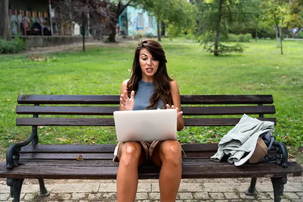Young freelance modern hipster woman working online in the park sitting on the bench having video call on web cam on computer. E-Commerce independent female small company owner marketing adviser