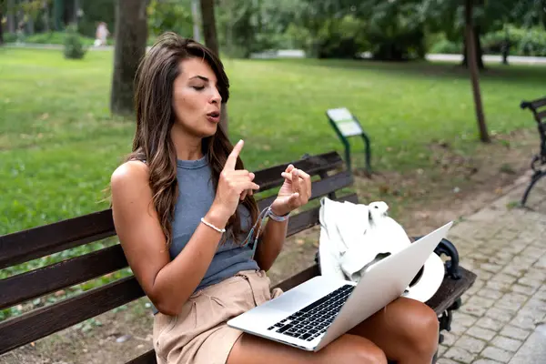 Young freelance modern hipster woman working online in the park sitting on the bench having video call on web cam on computer. E-Commerce independent female small company owner marketing adviser