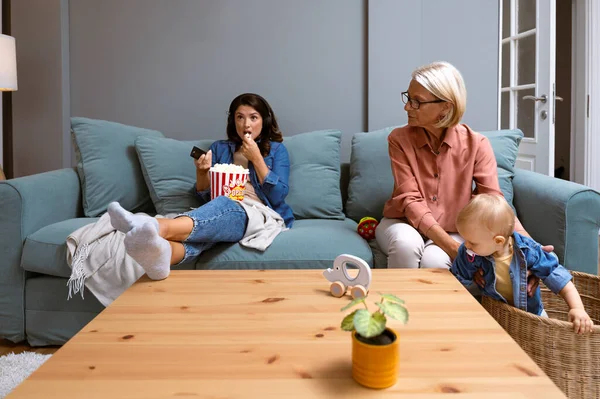Young irresponsible mother watch TV and eat popcorn while her mother child grandmother looks after the baby. Carefree mom pays no attention to her child who is being looked after by older babysitter