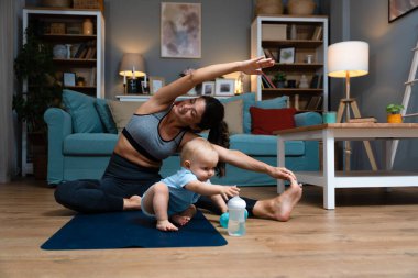 Genç ve mutlu bekar anne, yoga minderinde yoga yaparken bebeği dikkatsizce yerde oturuyor ve su şişesiyle oynuyor. Annem kaslarını esnetiyor.