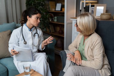 Mutlu genç bayan hemşire, evde bakım ziyaretinde gülümseyen yaşlı büyükanneyi desteklemeye yardım ediyor, bayan bakıcı doktor empati gösteriyor emekli hastayı ev hastanesindeki kanepede oturmaya teşvik ediyor.