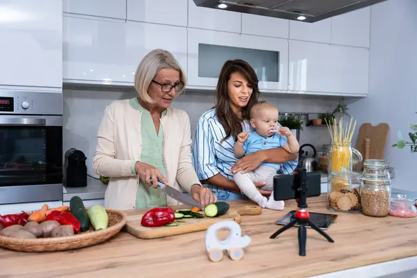 Grandmother mother and baby granddaughter cooking together in kitchen in apartment while mom making a funny faces for child and granny cutting vegetables on the kitchen countertop. Three generation