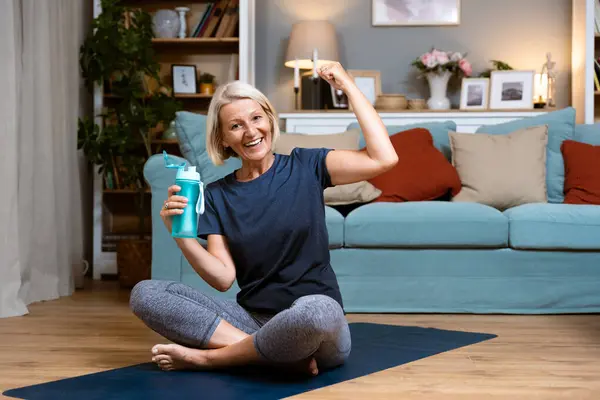 Senior woman exercising while sitting in lotus position. Active mature woman doing stretching exercise in living room at home. Fit lady stretching arms and back while sitting on yoga mat.