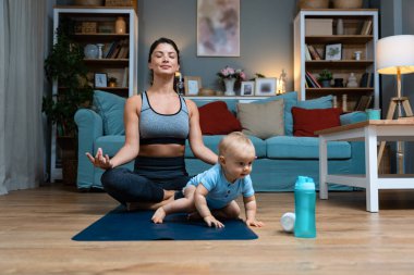 Genç ve mutlu bekar anne, yoga minderinde yoga yaparken bebeği dikkatsizce yerde oturuyor ve su şişesiyle oynuyor. Annem kaslarını esnetiyor.
