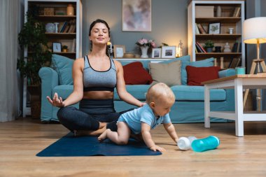Genç ve mutlu bekar anne, yoga minderinde yoga yaparken bebeği dikkatsizce yerde oturuyor ve su şişesiyle oynuyor. Annem kaslarını esnetiyor.