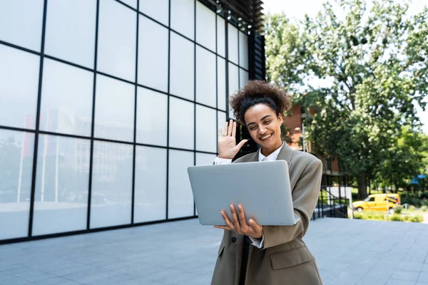 stock image Portrait of confident successful young curly woman, formally dressed, business woman standing with laptop outdoors against the background of the business center, looks at camera, smiling