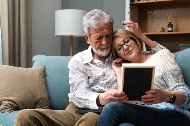 İki yaşlı çift, yaşlı çift, aile fotoğrafına bakıp gülümsüyor ve kanepede otururken sarılıyorlar. Çocuklar için anılar ve nostalji daha iyi bir yaşam için yurtdışına gitti.