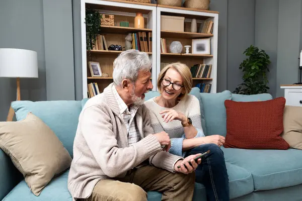 Happy senior old couple holding smartphone looking at cellphone screen laughing relaxing sit on sofa together, smiling elder mature grandparents family embracing having fun with mobile phone at home