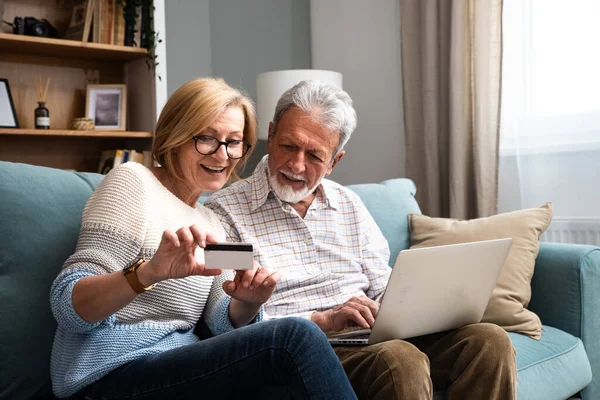 Cheerful old modern retired couple using credit card and laptop for online shopping, buying on Internet, choosing tour for vacation, making payment with bank application, financial service