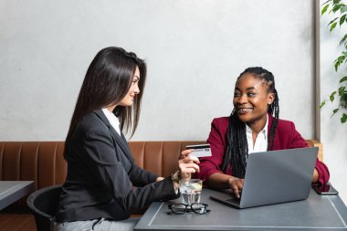 İki genç iş kadını küçük şirket sahibi kafeteryada oturmuş, online ödeme opsiyonları başvurusu için dizüstü bilgisayarlı banka kredi kartıyla yeni bir proje başlatıyorlar.