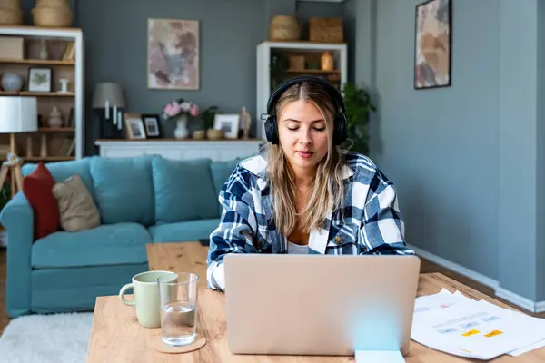 Happy woman wearing wireless headphones working on laptop at home office. Business female startup small company owner work on internet on computer.