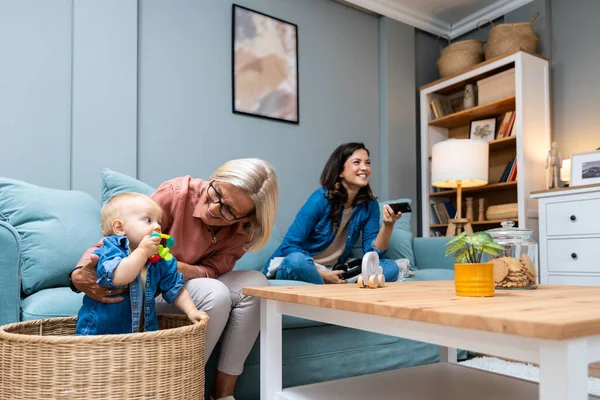 Young irresponsible mother watch TV and eat popcorn while her mother child grandmother looks after the baby. Carefree mom pays no attention to her child who is being looked after by older babysitter