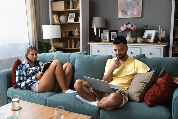 Happy young man working on sofa with laptop near woman with book lying on sofa in living room at home. Freelance male work on computer woman enjoying reading novel and resting. Couple goals concept