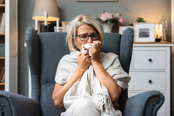 Senior woman sick and blowing nose at home from virus, allergy and illness on couch. Tired, tissue and elderly female person in a house lounge with sneeze from cold and flu on a sofa with allergies