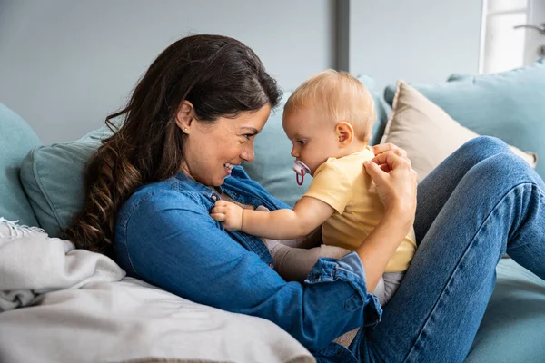 Aile, çocuk ve annelik kavramı. Mutlu gülümseyen genç anne, küçük bebeğe evde sarılıyor. Bir kadın ve yeni doğan oğlu ya da kızı annelerinin kucağında çocuk oyunu oynarken birbirlerine bağlanıyorlar.