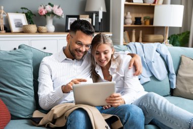 Resmi kıyafetlerle koltukta oturan heyecanlı genç çift tablet bilgisayarla online galibiyeti kutluyor, büyük bir anlaşma ya da iş başarısını evde kutluyor. Milenyum eşleri web mağazasında büyük indirimler yapıyorlar.
