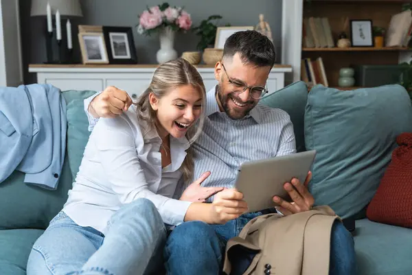Excited young couple sitting on couch in formal wear with tablet pc, celebrating online win, great deal or business success at home. Millennial spouses enjoying big sale in web store.