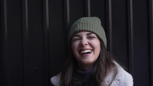 年轻快乐的微笑的女人站在室外 背对着黑墙 学生或自由职业者的快乐生活在外面 戴着冬衣 头戴羊毛帽子 穿着夹克 看着相机 — 图库视频影像