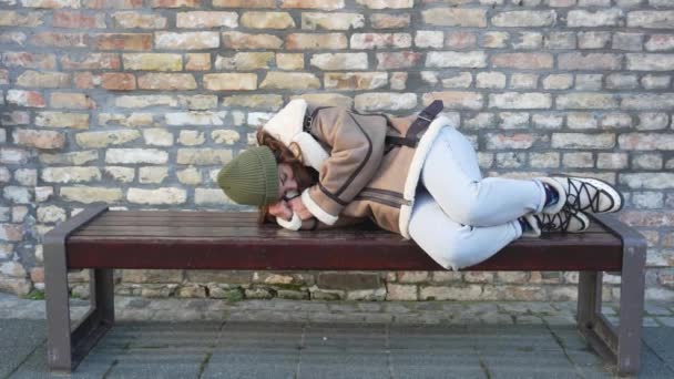路上でベンチで寝ているホームレスの少女 女性はバス停や鉄道駅で寒い天気を待っている列車やバスに乗り遅れた ホームレスと放棄された若者の問題 — ストック動画