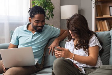 Cheerful millennial mixed couple spending time with laptop and book at home, happy young interracial lovers sitting on sofa with computer, freelance working or shopping online together and reading