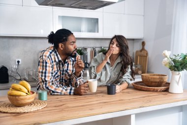 Multi-ethnic couple standing with tea cups in the kitchen and looks at each other with a love. African black man and a caucasian woman enjoy morning coffee together at home talking about life