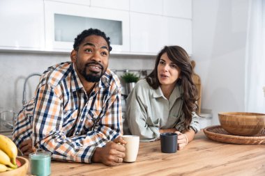 Multi-ethnic couple standing with tea cups in the kitchen and looks at each other with a love. African black man and a caucasian woman enjoy morning coffee together at home talking about life